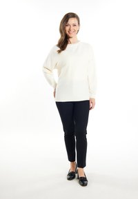 Losse crèmekleurige trui van tekstuurstof, met geribde manchetten en een bloemenpatroon rond de hals, gecombineerd met slim fit zwarte broek en donkere schoenen.