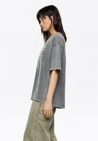 Camiseta gris de gran tamaño con mangas cortas, que presenta un estampado gráfico verde en la parte frontal. La tela tiene una textura suave y cómoda.