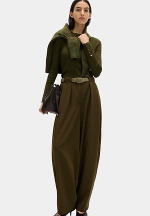 Frau trägt einen olivgrünen Pullover mit passendem Cardigan über den Schultern, weite olivgrüne Hose, Gürtel, schwarze Handtasche und High Heels.