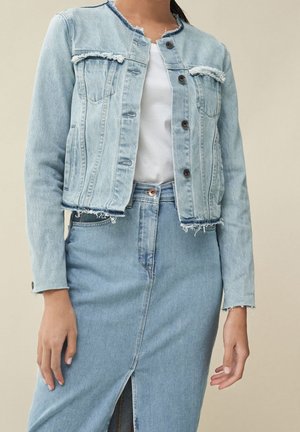 Femme portant une veste en denim bleu clair effilochée sur une chemise blanche et une jupe en denim assortie à taille haute avec une fente à l'avant.