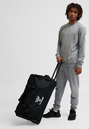 Svart reseväska med hjul, övre handtag, dragkedja och en logotyp. Tillverkad av hållbart tyg, med rektangulär form och slät textur.