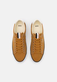 Par de zapatillas de lona marrón con suelas blancas, detalle de lengüeta a rayas negras y beige, y logo "BOSS" en las plantillas y los lados.