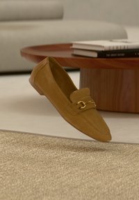 Mocassin en daim marron avec détail en m étal doré flottant au-dessus d'un tapis beige texturé, table basse ronde en bois avec des livres empilés en arrière-plan.