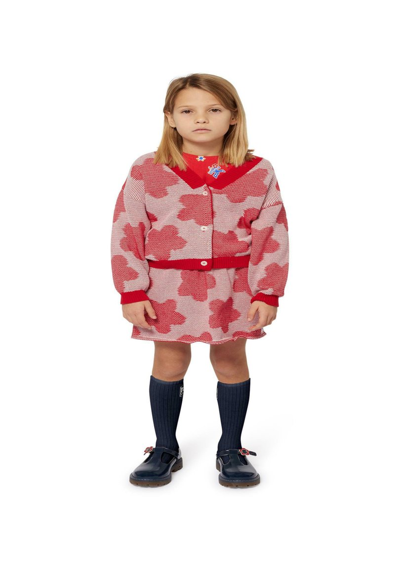 Robe rouge et blanche à motif floral, associée à un cardigan rouge et des chaussettes bleu marine jusqu'aux genoux. Comprend un badge sur le col et des chaussures foncées.