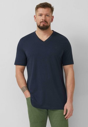 Mann mit Bart und Tätowierung am linken Unterarm, der ein marineblaues V-Ausschnitt-T-Shirt und grüne Hosen trägt, vor einem schlichten grauen Hintergrund steht.