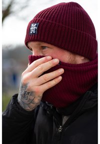 Burgunderfarbene Strickmütze mit geripptem Muster und Logo-Patch; passender gestrickter Halswärmer; getragen mit einer schwarzen Jacke.