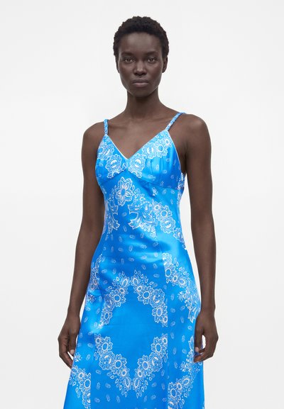 Femme mince portant une robe en satin bleu avec des motifs cachemire floraux blancs et des fines bretelles, debout sur fond blanc.