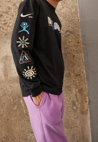 Svart långärmad tröja med färgglada grafiska tryck och logotyper på båda ärmarna, tillsammans med ljuslila sweatpants. Snyggt tyg.