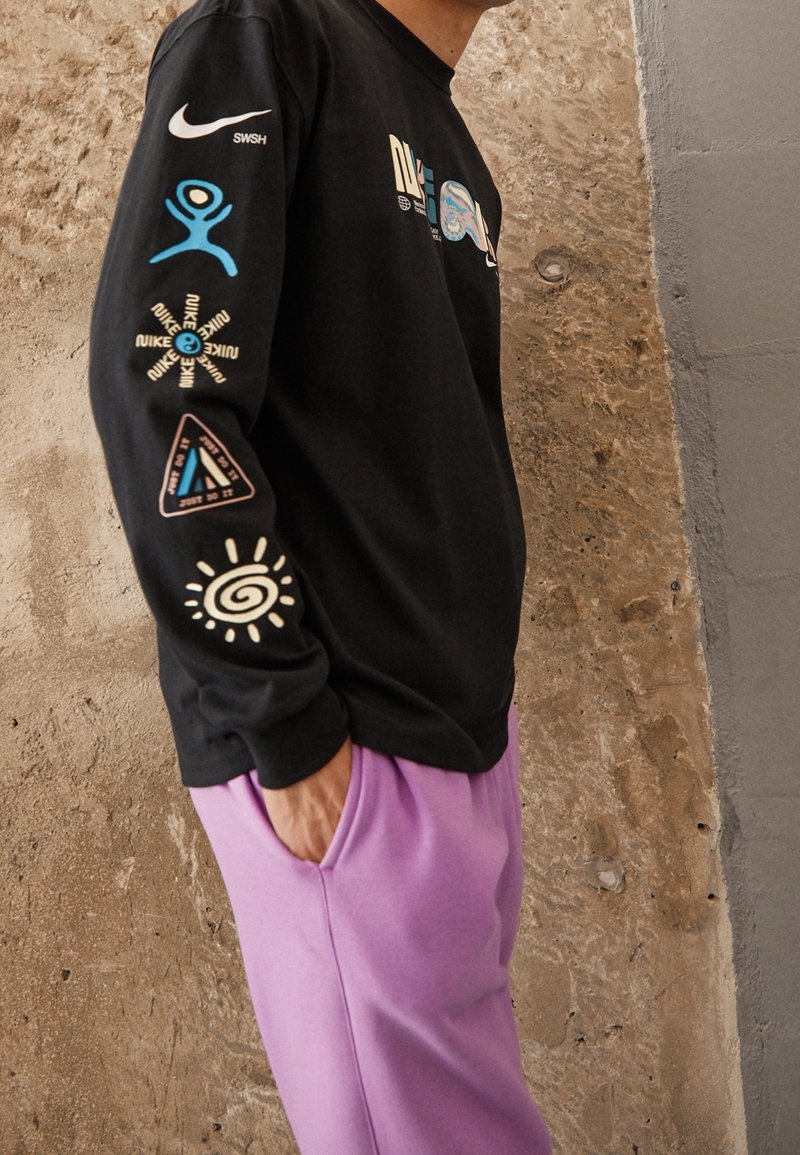 Svart långärmad tröja med färgglada grafiska tryck och logotyper på båda ärmarna, tillsammans med ljuslila sweatpants. Snyggt tyg.