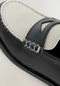 Mocassin en cuir noir et blanc avec un design cousu, présentant un accent découpé et une semelle texturée. Finition lisse sur le dessus.