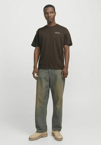 Brown cotton T-shirt with crew neck and short sleeves, featuring a small white print. Paired with loose, faded blue jeans and beige boots.