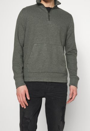 Homme portant un pull à fermeture éclair quart vert foncé avec poche kangourou à l'avant et jean noir usé sur un fond uni.