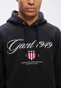 Bărbat îmbrăcat cu un hanorac negru pe care se află textul "Gant 1949", un emblema în culorile roșu, alb și albastru, și textul "American Sportswear Connecticut" în partea de jos.