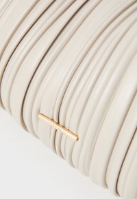 Vaalea nahkalaukku, jossa on uritett design. Varustettu erottuvalla kultaisella metalliosalla ja vaakasuoralla brändilevyllä. Suora pinta.
