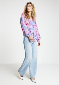 Transparente rosa Bluse mit blauen und violetten Blumenmustern, geraffter Vorderseite, kombiniert mit hellblauen Jeans mit weitem Bein und weißen spitzen Absätzen.