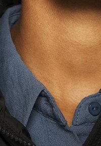 Effen button-up shirt van blauwe stof met een gestructureerde weving, gedeeltelijk bedekt door een zwart jasje. Heeft een zichtbare knop en kraagdetail.
