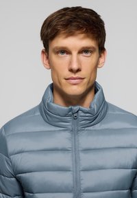 Ljusblå puffjacka med hög krage, med horisontell quiltad mönstring, framsida med dragkedja och en slät, glänsande yta.