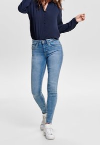 Jeans skinny blu in denim, con vita media, design a cinque tasche e un leggero sbiadimento. Abbinati a una blusa blu navy e a delle sneakers bianche.