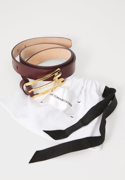 Curea din piele burgundy cu cataramă aurie, însoțită de un sac de praf din material alb, cu cravate negre și o etichetă pe care scrie "Victoria Beckham".