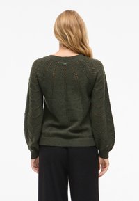 Γυναίκα με μακριά ξανθά μαλλιά, φορώντας ένα σκούρο πράσινο πλεκτό πουλόβερ με ανοιχτό σχέδιο και μαύρα παντελόνια, που στέκει στοργικά απευθυνόμενη προς τα πίσω.