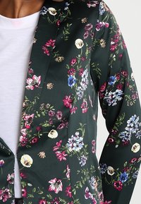 Marynarka w kwiaty w ciemnozielonym materiale, z różnorodnymi kolorowymi kwiatami, w tym różowymi, niebieskimi i białymi. Klasyczny krój kołnierza.
