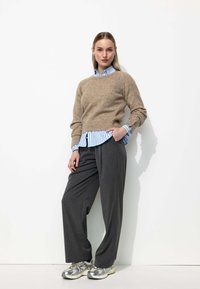 Un pull en laine beige sur une chemise à rayures bleues, associé à un pantalon large gris et des baskets argentées, sur fond blanc.