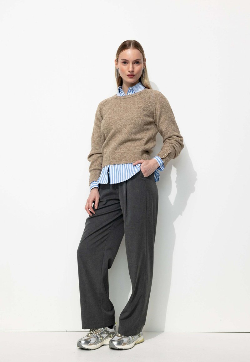 Un pull en laine beige sur une chemise à rayures bleues, associé à un pantalon large gris et des baskets argentées, sur fond blanc.