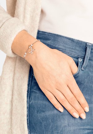 Silberner Gliederarmband mit einem X-förmigen Anhänger und klaren Steinen, getragen am Handgelenk, kombiniert mit blauem Denim und einem leichten Pullover.