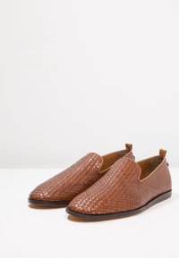 Chaussures en cuir tressé marron à enfiler, avec un bout pointu, une semelle plate et des côtés ouverts, présentant une texture lisse et des coutures minimales.