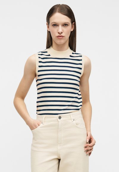 Γυναίκα με ίσια καστανά μαλλιά φοράει αμάνικο μπλουζάκι με ριγέ μοτίβο σε ναυτικό μπλε και λευκό, το οποίο είναι μπα tucked μέσα σε ανοιχτό μπεζ παντελόνι με υψηλή μέση, με το ένα χέρι στην τσέπη.