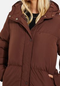 Chaqueta acolchada marrón con capucha, que cuenta con un cierre frontal de cremallera y botones, bolsillos laterales y una textura suave y lisa.