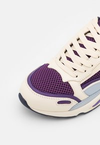 Detailní záběr na špičku tenisky s fialovou síťovinou, krémovými koženými překryvy, krémovými tkaničkami a světle modrými a fialovými akcenty na bílém pozadí.