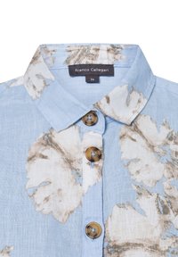 Lichtblauwe, opknopbare blouse met een bloemenprint in beige en wit. Voorzien van een klassieke kraag en bruine knoopaccenten. Maattag zichtbaar.