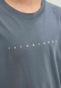Camiseta de algodón gris con cuello redondo y un logo impreso en blanco que dice "JACK & JONES" en el pecho. Textura suave.