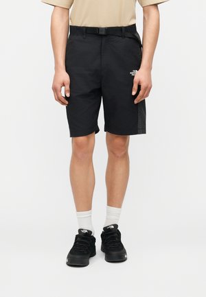 Άτομο που φοράει μαύρα σορτς The North Face με ενσωματωμένη ζώνη, λευκές κάλτσες και μαύρα αθλητικά παπούτσια The North Face, στέκεται μπροστά σε απλό φόντο.