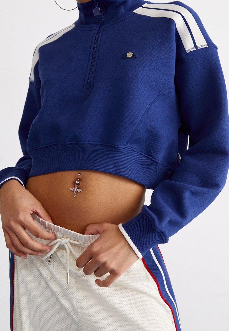 Felpa blu navy a maniche lunghe con collo a zip, strisce bianche sulle spalle e polsini a costine. Abbinata a pantaloni in rete bianchi con dettagli rossi.