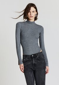 Junge Frau mit schulterlangem braunem Haar trägt silberne Ohrringe, einen grauen gerippten Rollkragenpullover und schwarze hoch geschnittene Jeans und posiert.