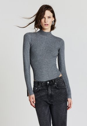 Una giovane donna con capelli castani alla spalla indossa orecchini in argento, un maglione grigio a coste con maniche lunghe e jeans neri a vita alta, in posa.