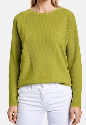 Femme portant un pull en maille vert à manches longues accompagné d’un pantalon blanc, montrée du cou aux hanches sur un fond uni.