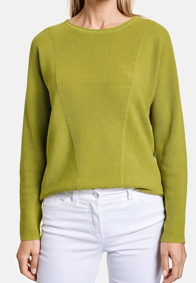 Femme portant un pull en maille vert à manches longues accompagné d’un pantalon blanc, montrée du cou aux hanches sur un fond uni.