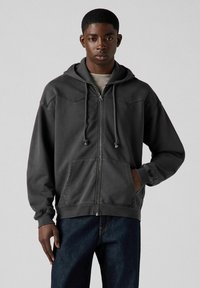 Sudadera gris con cremallera, capucha ajustable, bolsillos frontales y puños acanalados. Fabricada en tela suave con un corte relajado y sin patrones visibles.