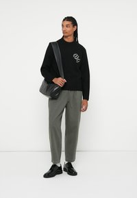 Svart strikkegenser med brodert logo, olivengrønne bukser, svarte lærsko og en crossbody-veske med glatt overflate og avslappet passform.