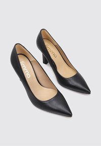Escarpins noirs en cuir avec un bout pointu, un petit talon bloc et une texture lisse. L'intérieur présente une doublure beige clair. Accentué par le nom de la marque.