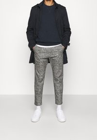 Mörkblå trenchcoat över en blå tröja, kombinerad med ljusgrå mönstrade byxor och vita sneakers med texturerad yta.