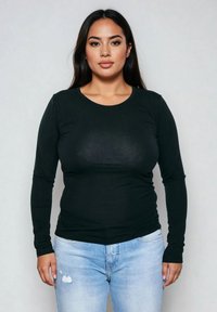 Noor naine pikkade tumedate juustega, seljas must pika varrukaga särk ja helesinised rebenditega teksad, seisab heleda halli tausta ees.