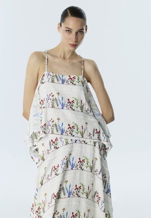 Frau mit dunklen Haaren, die ein weißes ärmelloses Kleid mit mehrfarbigen Blumenmustern vor einem schlichten hellen Hintergrund trägt.