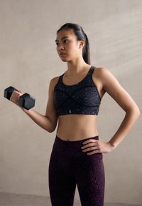 Juoda leopardų rašto sportinė pasaitė su plačiomis petnešomis, derinama su tekstūruotomis tamsiai purpurinėmis leggins. Asmuo laiko juodą hantelį.
