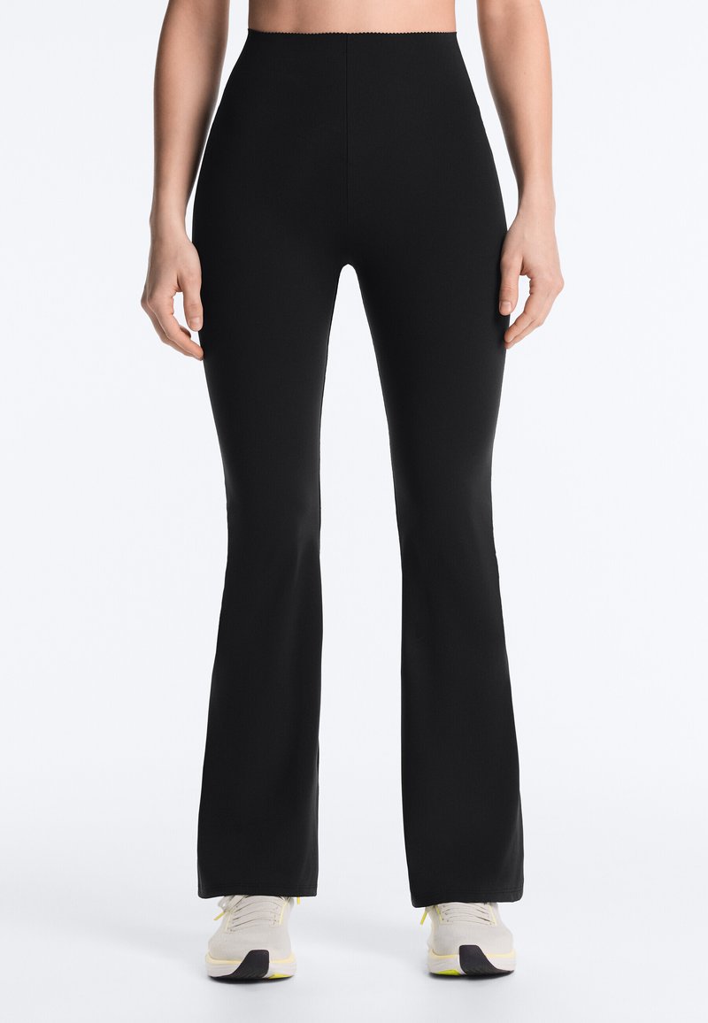 Woman wearing black high-waist, flared pants and white sneakers, standing with arms relaxed by sides against a plain background.