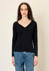 Ženska z kodrastimi lasmi, oblečena v črno majico z dolgimi rokavi in V-izrezom ter modre kavbojke, stoji pred enostavnim svetlim ozadjem.