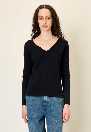 Žena s kovrčavom kosom u crnoj majici dugih rukava s V-izrezom i plavim trapericama, stoji na jednostavnoj svijetloj pozadini.